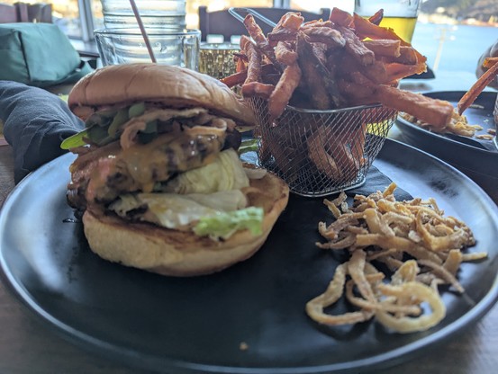 Cheeseburger  featuring arugula, iceberg lettuce, halloumi, crispy onions and cheddar cheese with A tiny fry basket of sweet potato fries and crispy onions on a black plate.  No side of sweet-hot chili sauce is visible anywhere.